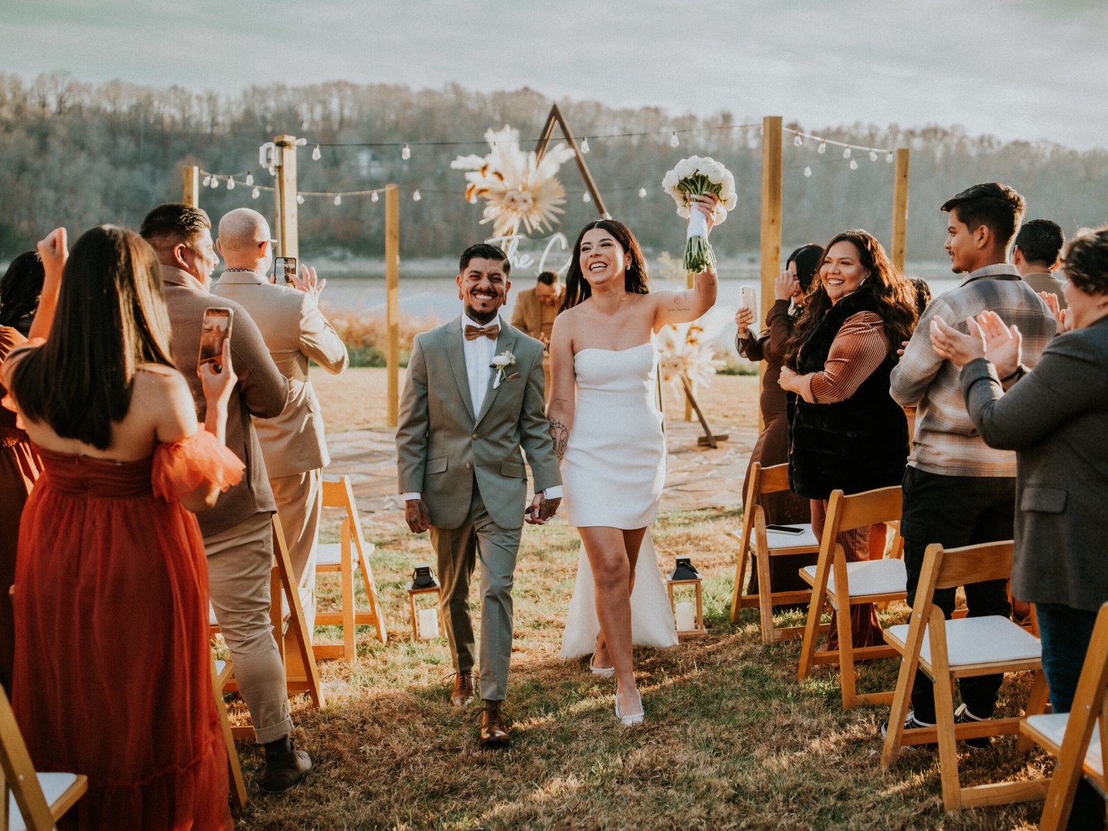 Newlywed couple holding hands and smiling as they exit their outdoor lakefront ceremony in Rogers, Arkansas.