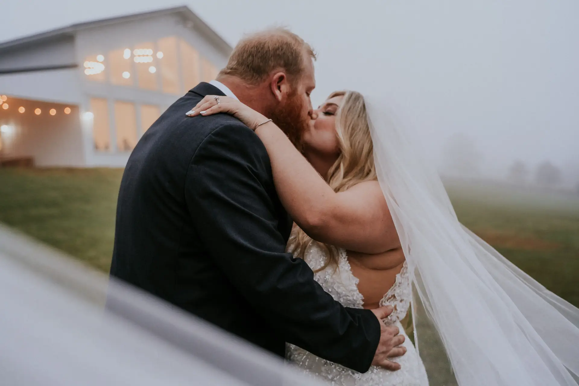 Newlywed couple sharing a kiss outdoors on a chilly, foggy day beside their wedding venue.