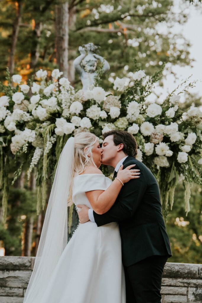 Bride and groom kissing in front of a white floral ceremony arch