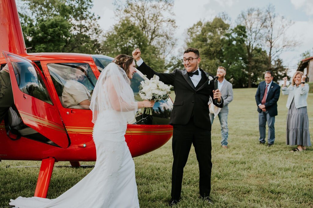 Bride and groom cheering beside a red helicopter on the meadow at Holland Barn in Highfill, Arkansas