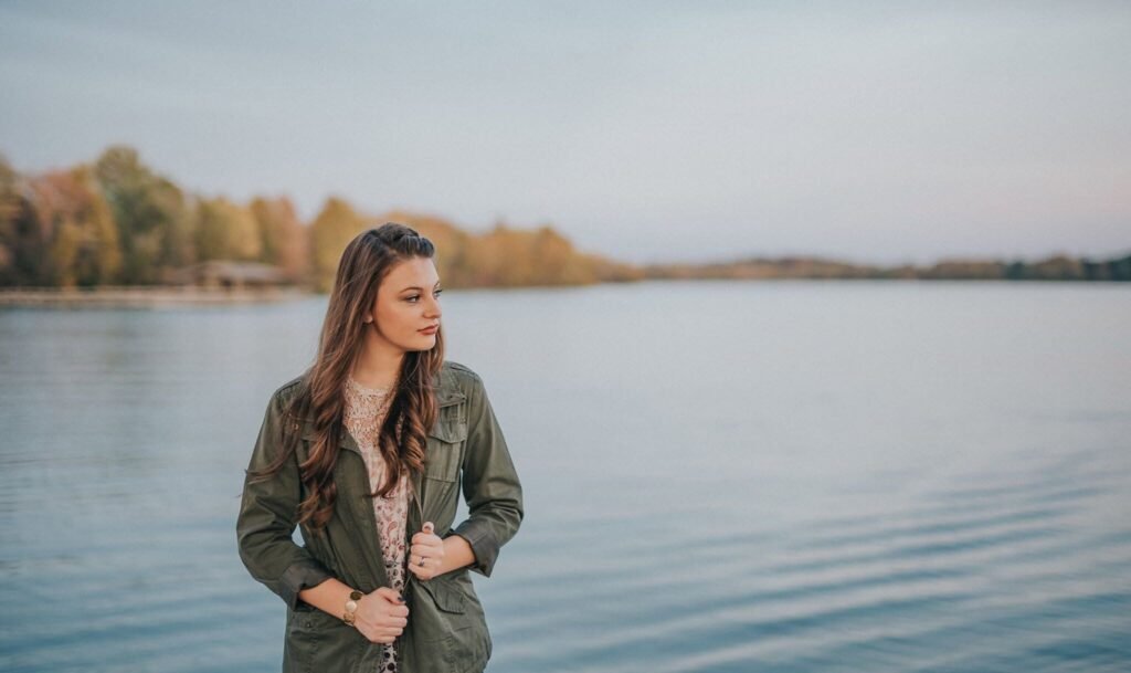 Senior girl in an olive jacket standing by the lake at sunset, NWA senior pictures
