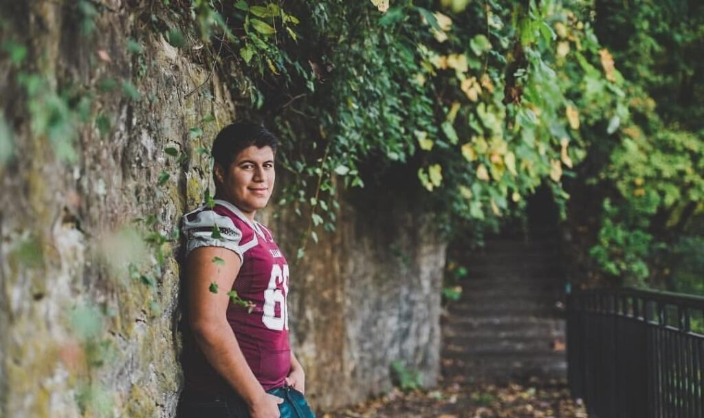 Senior football player in a maroon jersey leaning on a stone wall, NWA senior pictures