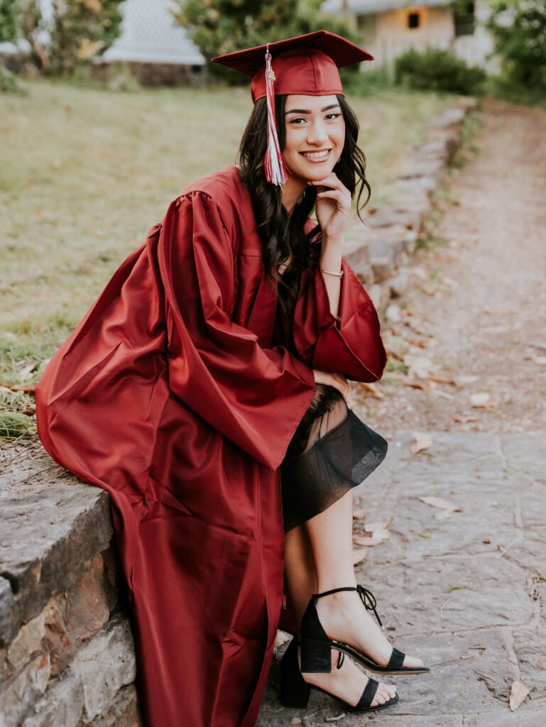 High school senior in a red cap and gown sitting on a stone wall