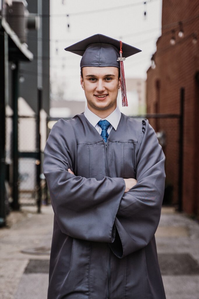 Senior guy in a gray cap and gown with arms crossed in a downtown alley, NWA graduation pictures
