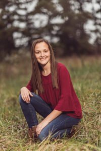 Senior girl in a red top sitting in tall grass at Gulley Park in Fayetteville, Arkansas