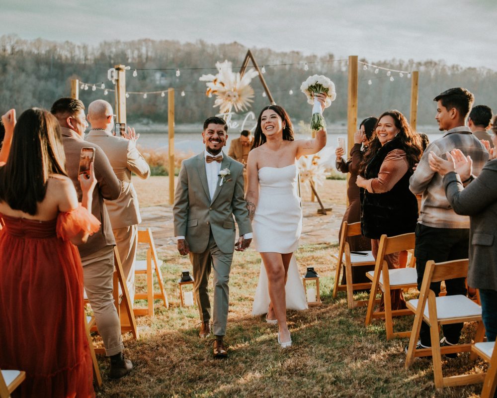 Newlywed couple holding hands and smiling as they exit their outdoor lakefront ceremony in Rogers, Arkansas.