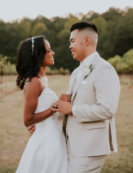 Bride and groom facing each other, holding hands in front of a vineyard, lost in each other's eyes.