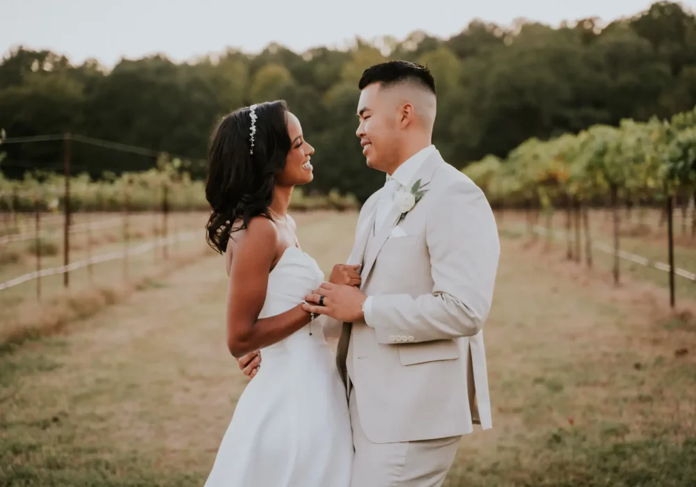Bride and groom facing each other, holding hands in front of a vineyard, lost in each other's eyes.