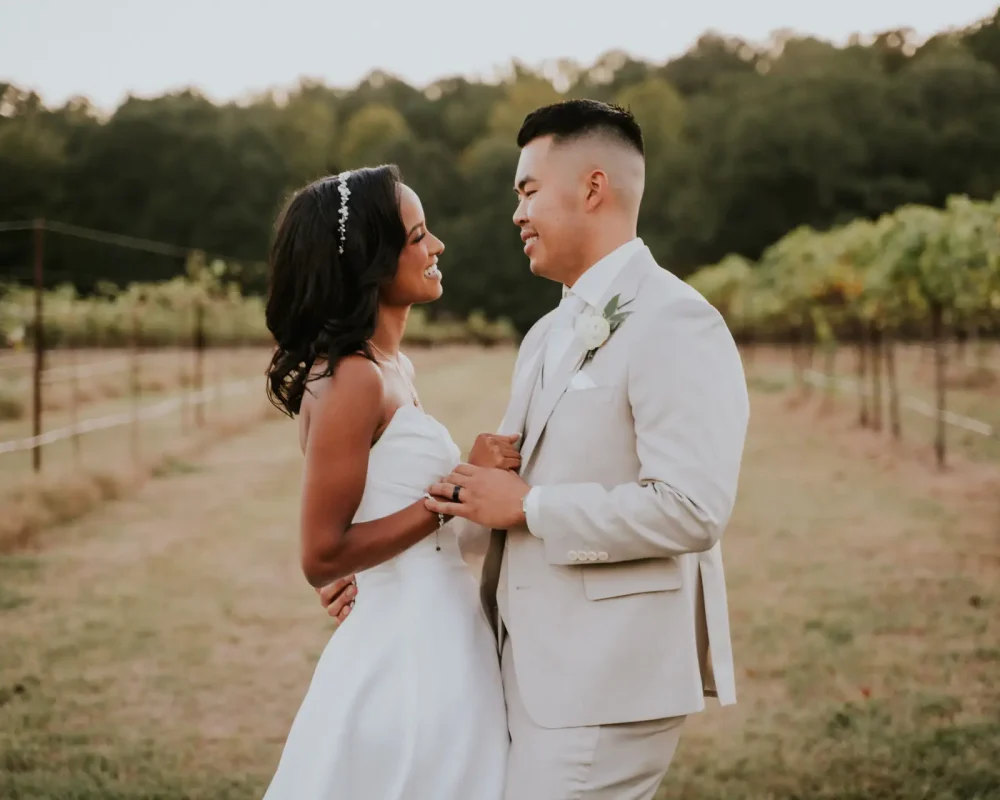 Bride and groom facing each other, holding hands in front of a vineyard, lost in each other's eyes.