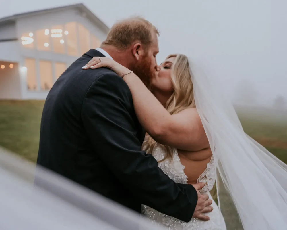 Newlywed couple sharing a kiss outdoors on a chilly, foggy day beside their wedding venue.